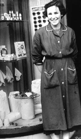Photograph of a woman wearing an overall of a dark colour with white piping and buttons, standing in front of part of a shop window containing a display of nail varnish, sun glasses, face flannels and face cream; she has been identified as Miss Williams in Haswell