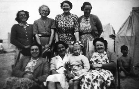 Photograph of four women standing behind three women sitting on deckchairs with a small girl, aged approximately six years, sitting on the lap of the woman in the middle of the three; the indistinct figure of a small boy can be seen to the right of the women sitting; behind the group are what appear to be wind breaks; the women are dressed in summer dresses and cardigans; they have been identified as members of Haswell Women's Institute on an outing to Tynemouth