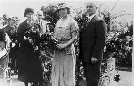 Photograph showing a woman dressed in a short-sleeved frock and a hat, standing holding flowers; another woman, wearing what appears to be a mackintosh and uniform hat, also holding flowers, is standing on the left of her; on the right of her is a man in a suit and tie; behind the woman in the mackintosh is the head and shoulders of another man in suit and tie; on the left of the photograph are part of three women with a perambulator; behind the group is display of flowers, almost as if it were in a glass house; the photograph has been identified as Garden Party at Tut Hill House, Haswell