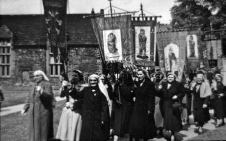 Photograph showing a procession of women wearing overcoats and cloths on thier heads walking with banners with saints on them; behind them are stone buildings with mullion windows; thay have been identified as Haswell Mothers' Union at Durham Cathedral
