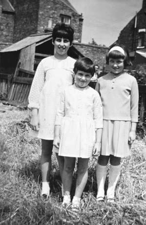 Photograph of three young girls, aged between six years and ten years, dressed in light-coloured dresses, standing on grass with the backs of houses and outhouses behind them; they have been identified as Anniversary Sunday, Haswell