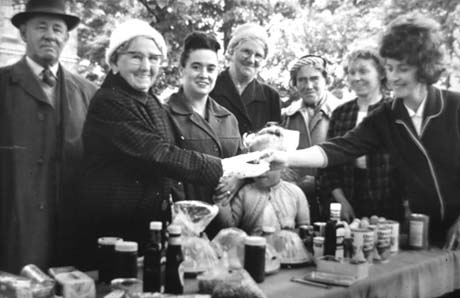 Photograph showing six women and one man, wearing overcoats, standing in front of a table on which bottles, tins, and bags of food are standing; another woman is handing some change to one of the women standing in front of the table; a small girl, aged approximately six years, is standing near the woman receiving change; the photograph has been described as Haswell Church Sale of Work