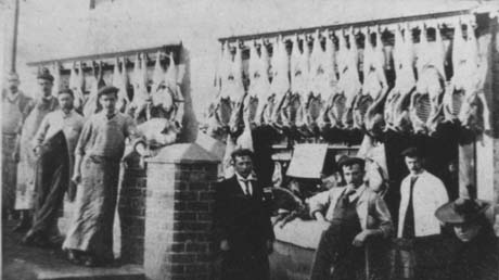Photograph showing six men in aprons, a man in suit, and the haed of a girl standing in front of a wall on which seventeen animal carcases are hanging; other indistinct items are lying on a what appears to be an open shop window near which two of the men are standing; the photograph has been identified as Co-Op Butcher, Haswell