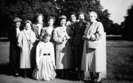 Photograph of eight women, dressed in overcoats, standing with trees and grass behind them; a ninth woman, wearing a dress and cardigan, is kneeling on the ground in front of the other women; they have been identified as members of the Haswell Womens' Institute on an outing