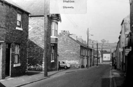 Photograph bearing a typed note reading: Station Street, Haswell, showing a street running away from the camera with two detached houses and a terrace of houses on the left; on the right are shops and houses; at the end of the street are buildings running across and open country beyond This photograph was submitted by Philip Soakell from the album 'History of Haswell and District', which was compiled by his grandfather Fred Soakell.