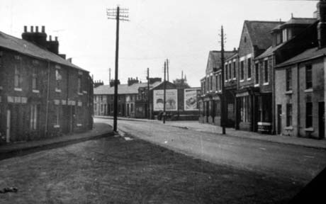 Photograph showing a street running away from the camera and another street, lined with terraced houses, running into the first; on the first street terraced houses, a large building with two gables and four shop windows can be seen indistinctly with bill boards just beyond it; the photograph has been identified as Co-Op and Front Street, Haswell This photograph was submitted by Philip Soakell from the album 'History of Haswell and District', which was compiled by his grandfather Fred Soakell.