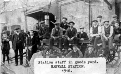 Photograph of twelve men and two boys, dressed in working suits and ties, sitting on a cart, with a brick building with a canopy over its entrance behind them; an indistinct figure can be seen standing near the entrance and a cart with boxes on it can be seen indistinctly in the distance; a note attached to the photograph describes it as Station Staff in goods yard. Haswell Station. 1916 This photograph was submitted by Philip Soakell from the album 'History of Haswell and District', which was compiled by his grandfather Fred Soakell.