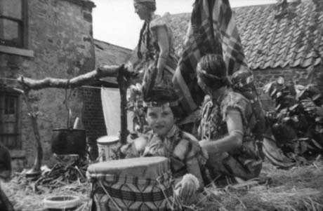 Photograph of a small child, aged approximately six years, dressed as a Red Indian with a drum in front of her; behind her are two girls, aged approximately thirteen years, also dressed as Red Indians; one is squatting behind the small child and the other is standing up near a pretend camp fire, over which a cooking pot is suspended; behind the children are the walls of houses built of rough stone and the children appear to be above ground level; they have been identified as being on a carnival float representing an Indian Reservation