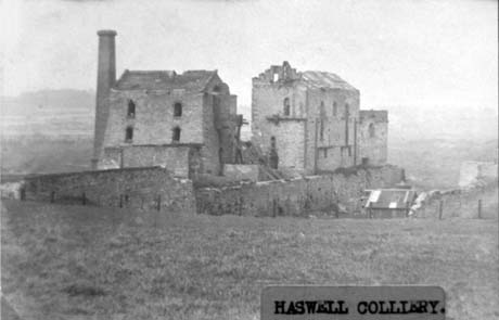 Photograph showing a large ruined brick building with a chimney next to it; at right angles to it is another ruined large brick building; behind and below them is a brick wall and, behind that, a shed; the buildings are in the middle of open countryside; they have been identified as Haswell Colliery This photograph was submitted by Philip Soakell from the album 'History of Haswell and District', which was compiled by his grandfather Fred Soakell.