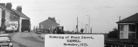 Photograph bearing a typed note reading: Widening of Front Street, Haswell. November, 1973. obscuring part of the photograph; a road running uphill at the left with terraced houses on its left side can be seen; there are cars parked in front of the houses and a lorry is proceeding up it away from the camera; on the right of the road there is an open space in which men and earth moving equipment can be seen indistinctly; a 1960s house can be seen on the right of the photograph This photograph was submitted by Philip Soakell from the album 'History of Haswell and District', which was compiled by his grandfather Fred Soakell.