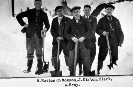 Photograph of four men, dressed in trousers, jackets and caps, standing in snow with an indistinct structure behind them; they are each holding a spade; they are accompanied by a man without a cap and wearing a collar and tie, who has been identified as the railway clerk;they have been identified as railway workers in Haswell and as follows: Left to Right: W. Hutton; C. Robson; J. Kirton; Clerk; A. Reay This photograph was submitted by Philip Soakell from the album 'History of Haswell and District', which was compiled by his grandfather Fred Soakell.