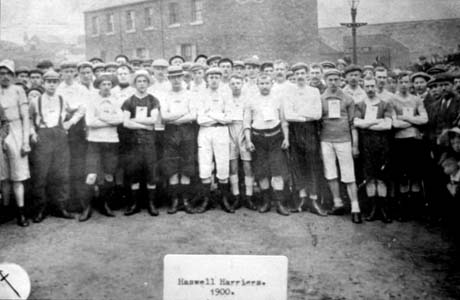 Photograph of a crowd of young men standing in a semi-circle with houses behind them; they are dressed in trousers, or short trousers, and vests, on which numbers are pinned; they are described in a typed note on the photograph as Haswell Harriers. 1900. This photograph was submitted by Philip Soakell from the album 'History of Haswell and District', which was compiled by his grandfather Fred Soakell.