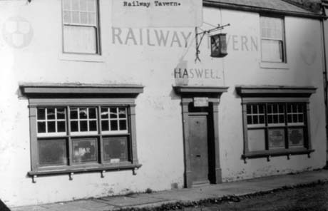 Photograph showing the exterior of a building with two windows with the word Bar on one and a door between the windows; above the door is a hanging sign that cannot be seen in detail; the words Railway Tavern Haswell are written on the wall This photograph was submitted by Philip Soakell from the album 'History of Haswell and District', which was compiled by his grandfather Fred Soakell.