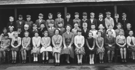Photograph showing forty children, aged approximately nine years, posed in three rows outside a covered way; a woman is sitting in the middle of the front row, presumably their teacher; they have been identified as Junior School Class J3, Haswell