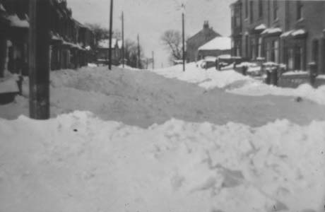 Photograph showing two rows of houses with bay windows on a gently rising slope; in between them is deep snow and the roofs and ledges of the houses are covered with snow; the photograph has been identified as Top of East Villas - Snow Storm, Haswell