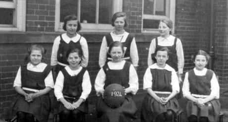 Photograph of eight girls, aged approximately thirteen years, wearing gym slips, po ed in two rows outside a brick building; thay have been identified as First Netball Team At County School, Haswell; a girl on the front row is holding a ball bearing the date 1924