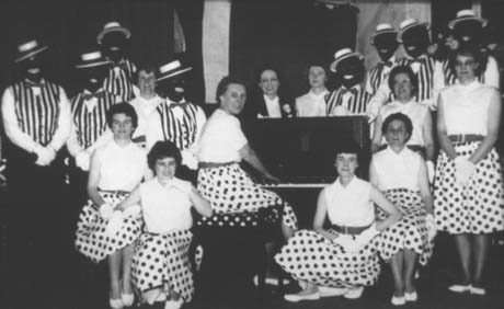 Photograph of nine women, wearing light-coloured blouses and spotted skirts, and eight men with blacked faces, wearing striped waistcoats and boaters, sitting and standing round a piano at which one of the women is sitting; behind the piano is another woman dressed in male evening clothes of dinner jacket and white tie; they have been identified as Black and White Minstrels, Haswell
