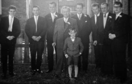Photograph of eight men standing against a low fence with brick buildings in the background; they are all wearing suits and ties and some have buttonholes pinned to their coats; a boy, aged approximately eight years, and dressed in a suit with short trousers, is standing in front of them; they have been identified as taking part in The Johnson/Champley Wedding in Haswell