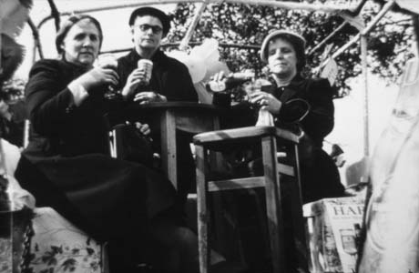 Photograph showing a woman sitting at the left of the photograph on an upholstered seat and holding a glass of beer; next to her is a man, wearing a dark beret and dark glasses, holding a glass and leaning on what may be a table; next to him is a second woman wearing a coat and hat and pouring beer from a bottle into a glass and sitting behind a stool; all three are in the open air with a structure of metal poles above them through which trees can be seen; they have been identified as Women's Group, Coronation Street, John Brazier English, Haswell