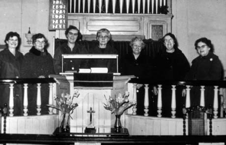 Photograph showing seven middle-aged women, wearing dark overcoats, standing with their backs to the organ and behind the balusters of the dais, in a church, identified as the Methodist Church in Haswell; a crucifix and two vases of flowers are standing in front of the balusters