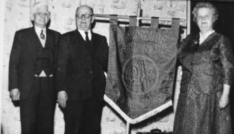 Photograph of two elderly men in dark suits standing on the left of a banner held by an elderly woman in a dark dress and jacket; the banner reads: Haswell Branch No. 1 Region OAP; they are standing against a wall covered in wallpaper