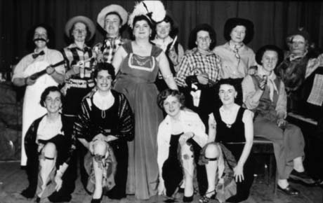 Photograph showing thirteen women, in costume, posed in two rows on a stage; the women on the back row are dressed as cowboys and the keeper of a saloon; one woman is sitting at a piano at the right of the picture; a woman in the middle is dressed as a saloon girl and four women on the front row are dressed as saloon girls and kneeling showing their legs and garters; they have been identified as members of Haswell Women's Institute