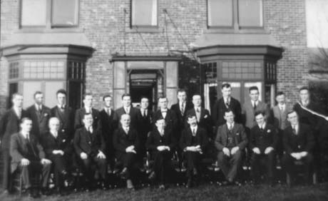 Photograph of twenty three men in suits and ties, posed in two rows, in front of a house with two bay windows and an imposing doorway; they have been identified as members of the St. John Ambulance Brigade in Haswell