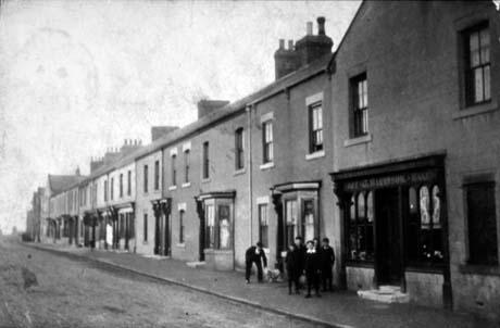 Photograph of a street, showing a terrace of houses with four or five fitted out as shops; two houses have bay windows and two or three have elaborate doorways; the details of the shops cannot be seen; five indistinct boys and an indistinct man and a dog are on the pavement; the street has been identified as Church Street, Haswell