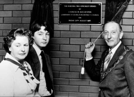Photograph of a man wearing a suit, tie and chain of office, posed pulling the cord of a curtain, which has covered a plaque recording the opening of a building; opposite him are middle-aged woman and a boy, aged approximately fifteen years; the plaque records the opening of the building, which has been identified as Seaham Leisure Centre, by Councillor Alex McAteer, Chairman of Easington District Council on Friday 29th August 1980