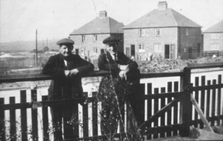 Photograph of two elderly men, wearing suits and caps, leaning on a garden fence looking at the camera; behind the men are a road, open ground and three semi-detached houses; the photograph has been described as Haswell Before Bungalows Are Built