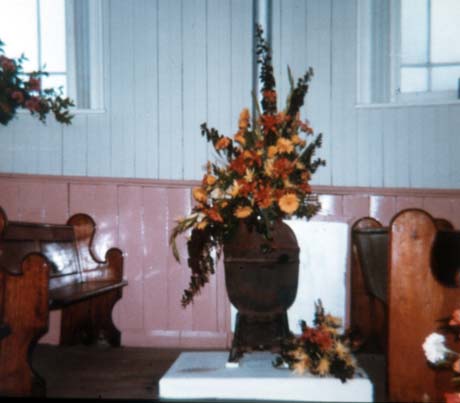 Photograph of an arrangement of orange and bronze flowers, displayed between two pews and in front of a wooden wall, painted blue and pink, in a church; the photograph has been described as Flower Festival in Chapel, Haswell