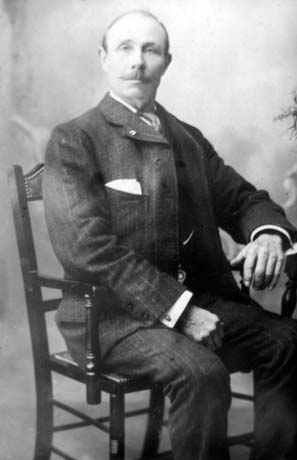 Photograph of a man sitting very straight on an upright chair, resting his left arm on a table; he is wearing a pinstriped suit, has a moustache, and has receding hair; he has been identified as Mr. Smith The Local Preacher, Haswell