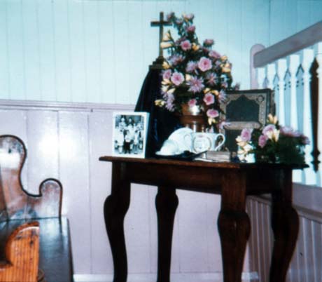 Photograph showing an arrangement of pink and yellow flowers on a table with a crucifix, a bible, a photograph, a commemorative mug, and a miner's helmet; the display is on a table near a pew with a blue and pink wall behind it; the display has been described as being in part of a Flower Festival in Haswell Plough Chapel; it is, most likely, part of the same display as that in0196