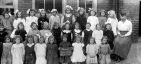 Photograph of twenty nine girls, aged approximately five years, posed in three rows outside a brick building, with a woman sitting at the right of the group; a child on the front row is holding a notice, which reads: Haswell Council Girls Standard 1 1919