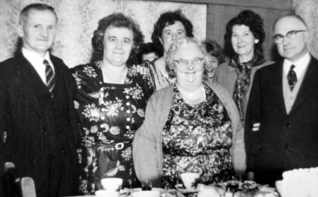 Photograph showing a middle-aged woman in a dress and cardigan and a middle-aged man in a suit, standing behind a table on which cups and saucers and other crockery are laid; behind them are a middle-aged man in a suit and three middle-aged women in dresses, and a young woman, standing against the wall of a room; they have been identified as Birthday Party with Mrs. Howie and Family