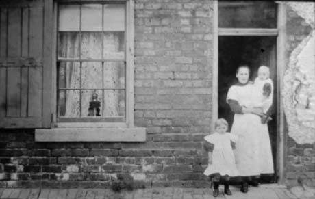 Photograph of a woman, wearing a long light-coloured pinafore over a dark garment, standing in the doorway of a brick house with a sash window at which there are net curtains and a shutter; she is carrying an infant, and a small child of approximately three years, wearing a pinafore and dark stockings and boots, is standing next to her; they have been identified as Mrs. Errington and Two Children in Haswell