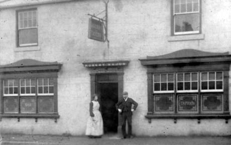 Photograph of the exterior of a public house showing two windows, with an elaborate wooden surround, on which the words Bar and Taproom can be seen; there is a hanging sign above the door which cannot be read, but the public house has been identified as The Railway Tavern in Haswell; a woman wearing an apron to her ankles and a man in a suit and cap are standing outside the doorway