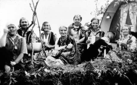 Photograph of seven women dressed as gypsies sitting on branches and leaves round a tripod, made of twigs, from which a cooking pot is suspended; one of the women is holding a doll and feeding it with a bottle; another is holding an accordion; at the right of the picture the front of a gypsy caravan can be seen; the photograph has been described as Haswell Women's Institute Gypsy Encampment