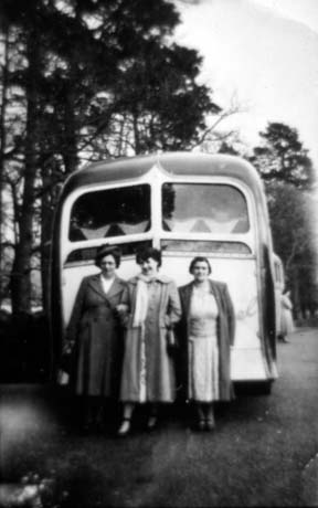 Photograph of three women in overcoats standing with the rear of a single-decker bus behind them; there are trees behind the bus; thay have been identified as Mrs. Vera Howie; Mrs. Wilkinson; Mrs. Morrow