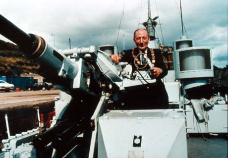 Photograph of a man wearing a suit and a chain of office, standing at the controls of a large gun on board a ship; the mast and equipment of the ship can be seen in the background; the man has been identified as Councillor A. McAteer, Chairman of Easington District Council