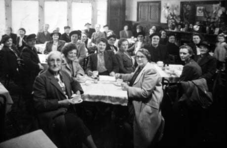 Photograph of approximately thirty five middle-aged women, one man and one child aged approximately three years, sitting at approximately eight tables in a room with large windows along one wall and a door and dresser along the other; they are wearing overcoats and suits; they have been described as Haswell Ladies' Outing