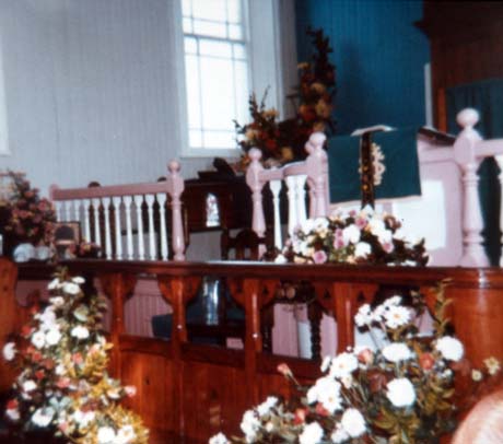 Photograph of four flower arrangements in front of the balusters of a dais and in front of a communion rail in what has been identified as Haswell Plough Chapel; the walls and a window of the chapel can be seen