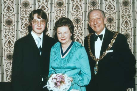 Photograph showing a man in evening dress and a chain of office, standing posed against elaborately patterned wallpaper; next to him is a woman wearing a turquoise evening dress and a ribbon of office, carrying a posy of flowers; next to her is a boy, in a suit and tie, aged approximately fifteen years; they have been identified as Councillor Alex McAteer, Mrs. Margaret McAteer, and their son, Alex McAteer
