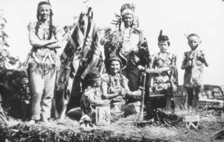 Photograph of three women and three children dressed as Red Indians; one woman, dressed as a squaw, and one woman dressed as a brave, are standing at the back of the group; one woman and one child are sitting near the camp fire and two children are standing to one side; they are most likely on a float at a carnival; thay have been identified as members of Haswell Women's Institute