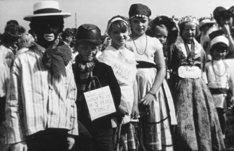 Photograph of a crowd of people, the front row of whom are children in fancy dress; they are aged approximately between ten and twelve years; one girl is dressed as Queen Elizabeth; another as a beauty queen; one boy is dressed as a coal miner and has a notice round his neck reading; What Will Happen To Me In 5 Years Time?; they have been identified as taking part in Haswell Plough Carnival