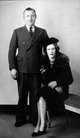 Photograph of a man, wearing a suit and tie, standing beside a woman who is sitting on a bench; she is wearing a dark hat, a dark coat with a fur collar, gloves and high-heeled court shoes; they have been identified as Ivy and Hiram Brass of Haswell