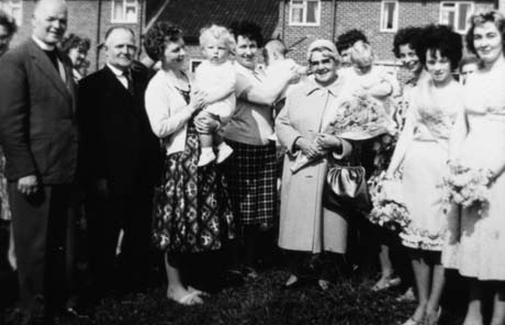 Photograph showing a clergyman and a man in a suit standing at the left of the picture; next to them are three young women holding infants; next to them is a middle-aged woman holding a bouquet of flowers; next to her are three young women holding bouquets of flowers; one of these has a sash across her chest; behind the group are the walls of post-Second-World-War houses; the photograph has been identified as Haswell Bonnie Babies Competition, and the clergyman as the Rev. Mr. Sparks and the other man as Councillor Yews