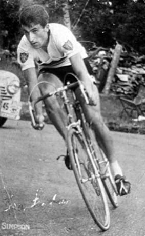 Photograph of a man in a shirt with Union Jack badges on its sleeves and dark shorts riding a racing bicycle towards the camera; part of the front of a car can be seen at the left of the picture; the surface of the road and woodland verge can be seen behind the cyclist; he has been identified as Tony Simpson International Racing Cyclist; the name Simpson is printed on the photograph