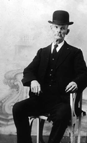 photograph,most likely taken in an artist's studio, of an elderly man sitting on a wooden chair with arms; he is wearing a Bowler hat; dark suit and waistcoat and a dark tie; he has been identified as Mr. Barass of Haswell