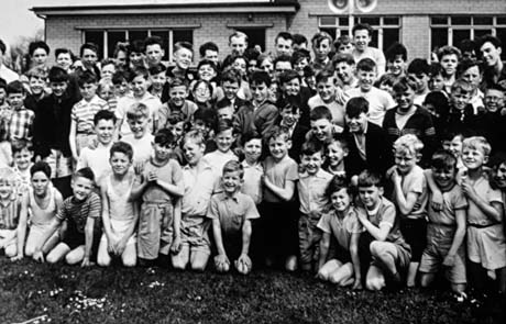 Photograph of a crowd of boys, aged between approximately eleven and fourteen years, posed with a low brick building behind them; some are wearing shorts and singlets and may have been taking part in sport of some kind; they have been identified as being in Haswell